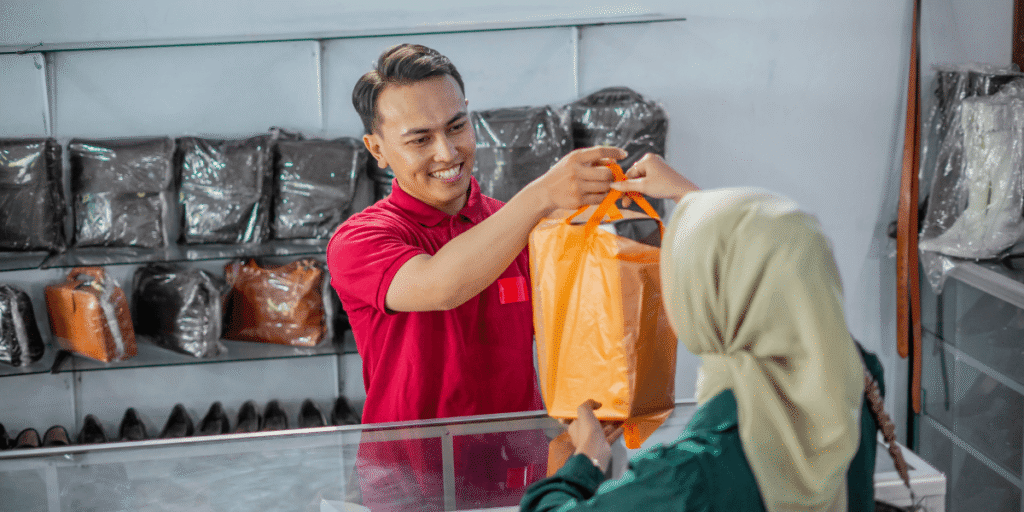 A smiling clerk in a red shirt hands a customer an orange shopping bag at a store counter, with bags and shoes displayed in the background.