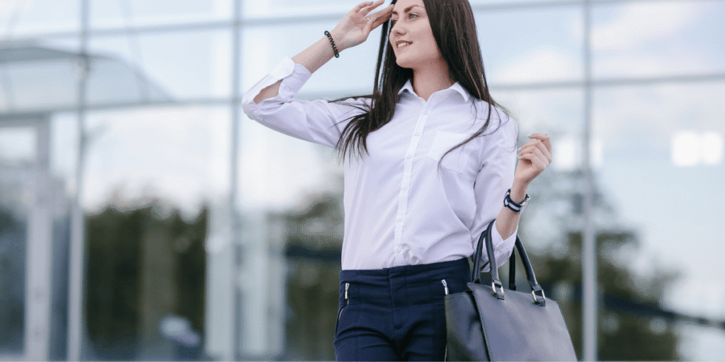 A woman in a white shirt and black pants holds a bag, standing confidently with a neutral expression.