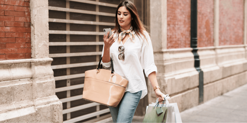 A woman walks down the street carrying shopping bags in both hands, smiling as she enjoys her day out.